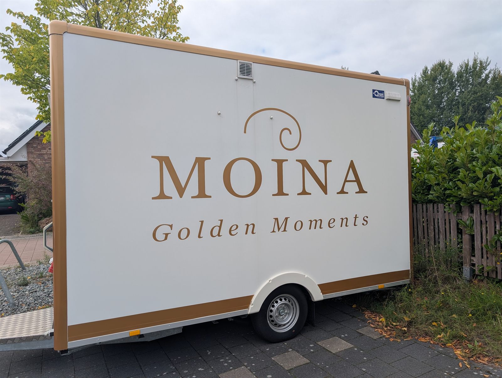 Der MOINA-Verkaufsanhänger von der Seite: weiß mit goldenen Kanten und goldenem Schriftzug „MOINA – Golden Moments“